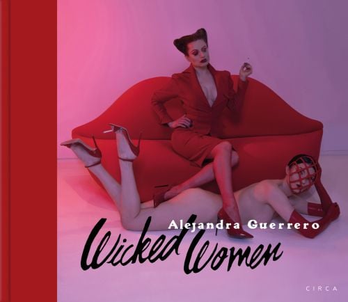 Woman in red suit sitting on Mae West lips sofa, with her red stilettos across a nude woman laying on floor, on cover of 'Alejandra Guerrero - Wicked Women', by Circa Press.