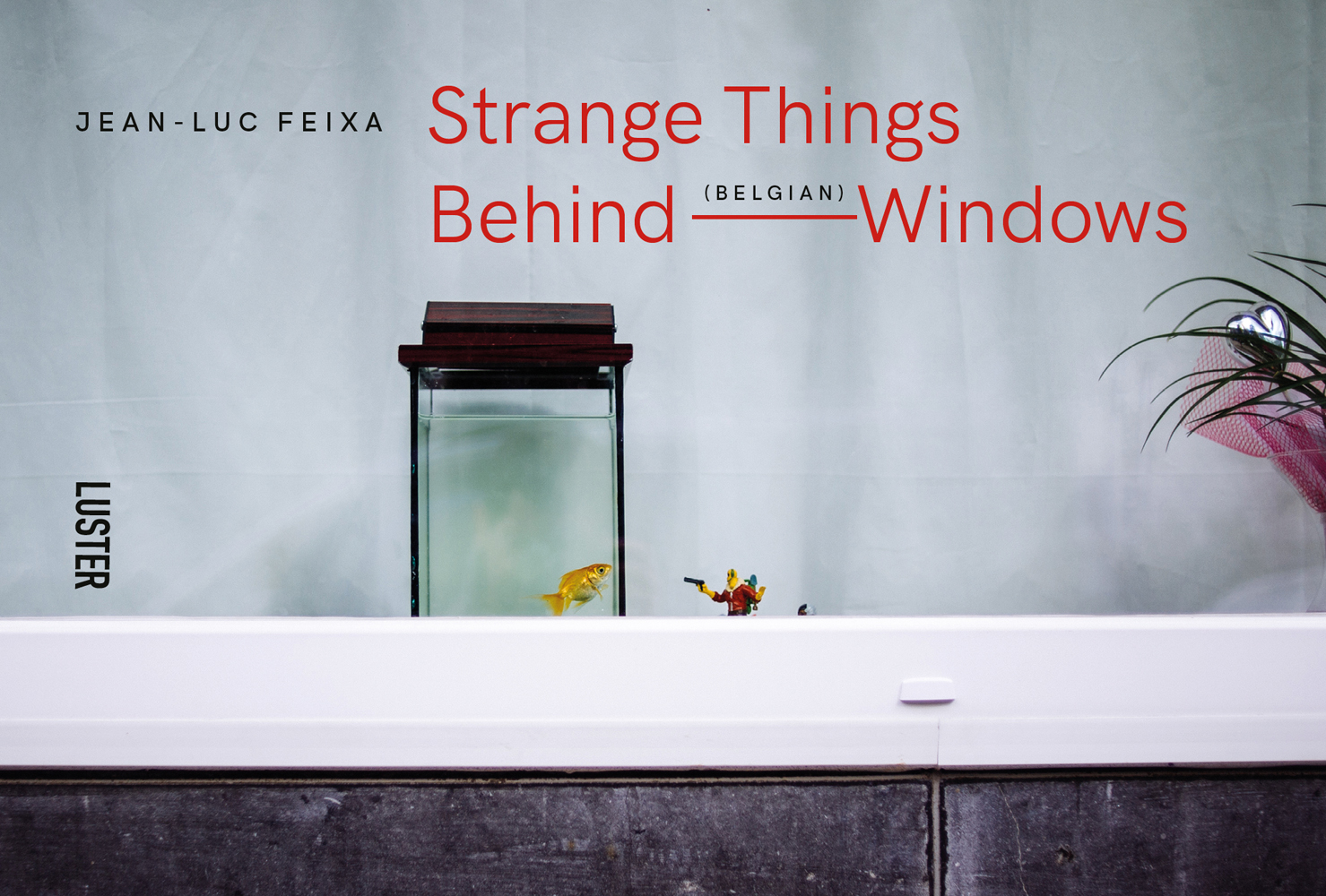 Small glass oblong fish tank with a goldfish, on windowsill, white curtain behind, Strange Things Behind Belgian Windows in orange and black font above.