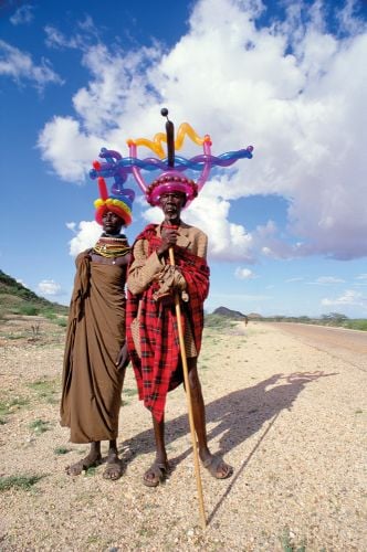 2 African men in traditional dress, one with stick, wearing crowns on their heads made from colourful inflatable balloons.