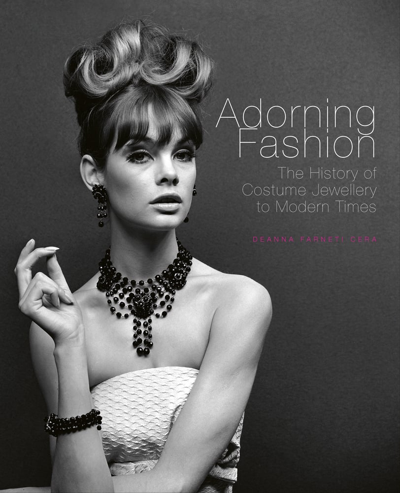 Half length black and white photo of a fashion model with an up-do wearing a beaded necklace and dress gazing nonchalantly at the camera with Adorning Fashion The History of Costume Jewellery to Modern Times in white font