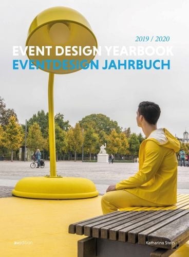 Man in yellow waterproofs sitting on bench, staring at gigantic yellow desk lamp, 2019/2020 EVENT DESIGN YEARBOOK in blue and white font above.