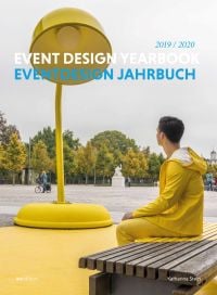 Man in yellow waterproofs sitting on bench, staring at gigantic yellow desk lamp, on cover of 'Event Design Yearbook 2019/2020', by Avedition Gmbh.