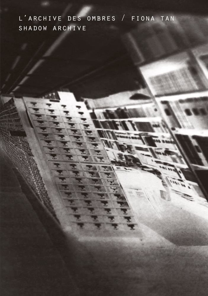 Black and white still of building structure, with ramp, L'ARCHIVE DES OMBRES / FIONA TAN SHADOW ARCHIVE in white font above.