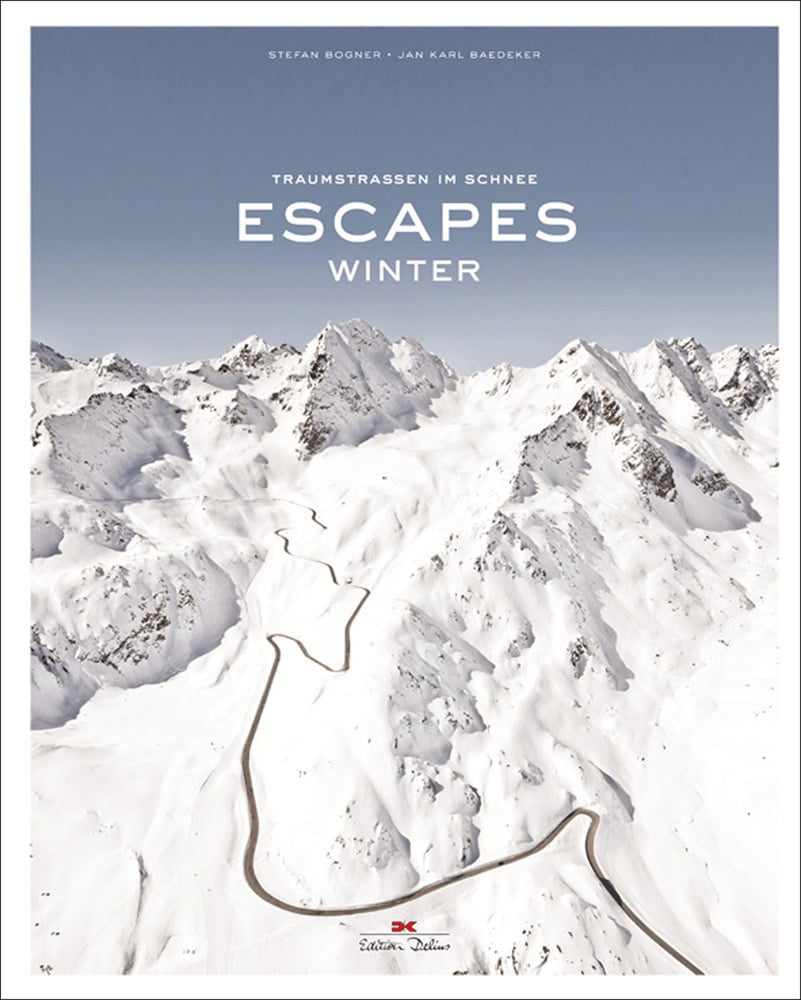 Aerial view of snow covered mountains with winding road, on cover of 'Escapes: Winter', by Delius Klasing.