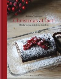 Christmas chocolate roulade, sprinkled with icing sugar, on glass dish, on cover of 'Christmas at Last! Holiday Recipes and Stories from Italy', by Guido Tommasi Editore.