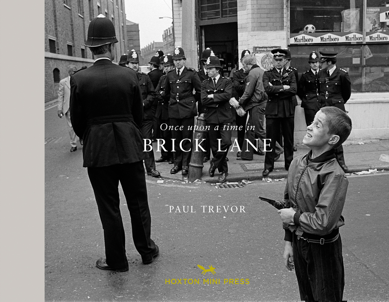 Line of policemen on street corner, young white child standing in front holding gun, pulling face, on landscape cover of 'Once Upon a Time in Brick Lane', by Hoxton Mini Press.