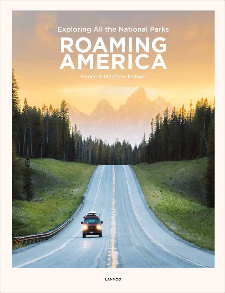 Campervan on straight road with mountainous landscape behind and orange sky, on cover of 'Roaming America, Exploring All the National Parks', by Lannoo Publishers.