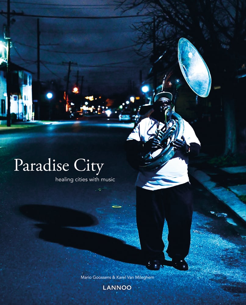 Black musician playing a sousaphone while walking the streets at night, Paradise City in white font to centre left.