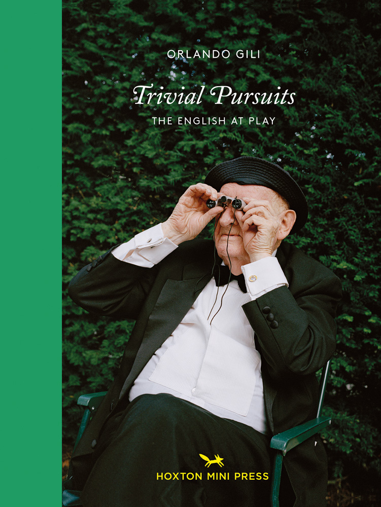 Older white man sitting in garden chair looking through small binoculars, with green yew hedge behind, on cover of 'Trivial Pursuits, The English at Play', by Hoxton Mini Press.