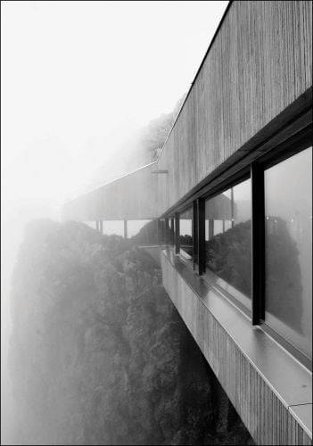 Misty black and white shot of side of modern, flat roofed building, sheer drop with trees below.