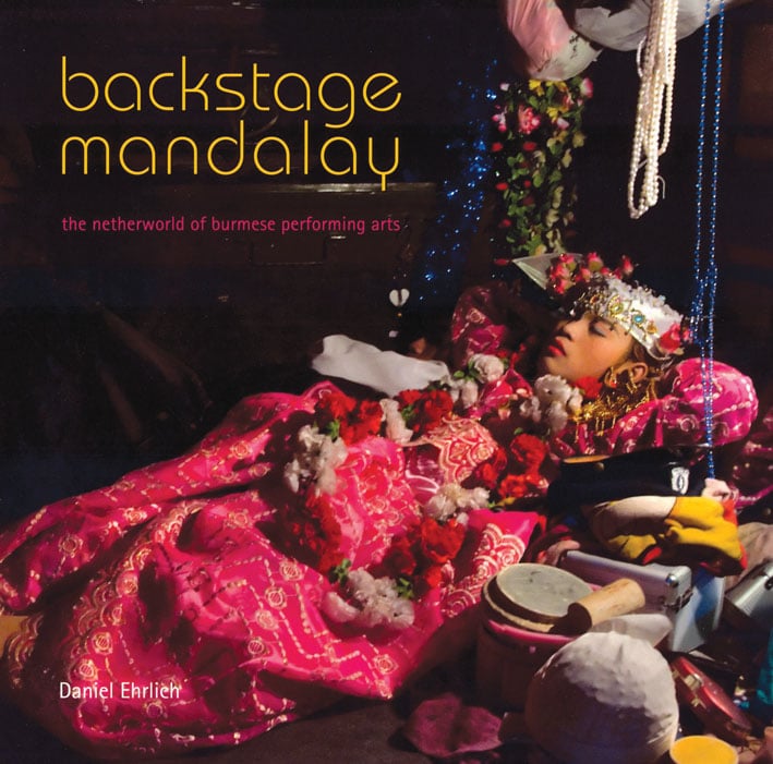 Performing arts theatre scene with young Burmese girl wrapped in pink cloth, flowers on top, on cover of 'Backstage Mandalay', by River Books.