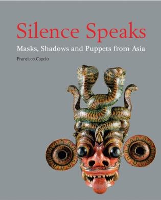 Exhibition photograph of carved Sri Lankan mask with projecting tongue in pierced jaws and carved teeth, on cover of 'Silence Speaks', by River Books.