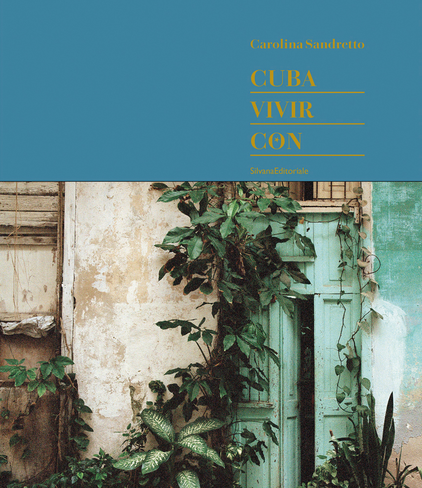 Front of old Cuban house, tropical green foliage growing up around door frame, CUBA VIVIR CON in gold font on blue banner above.
