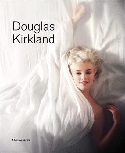 Marilyn Monroe in playful pose, laying beneath a white sheet on bed, Douglas Kirkland in black font to upper left.