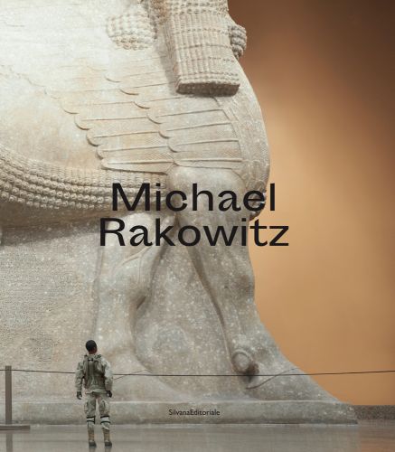 Lower portion of giant lamassu in exhibition space, black man in military uniform looking up, Michael Rakowitz in black font to centre.