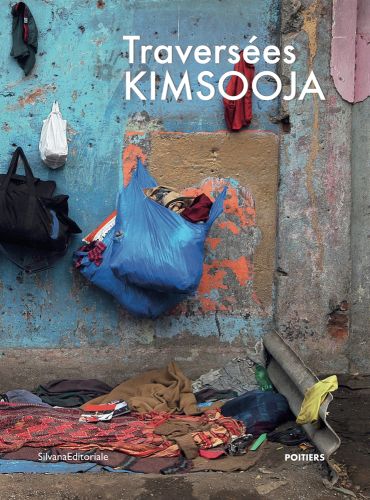 2 blue plastic bags filled with clothes hang on a nail in a concrete wall, above blankets on floor, Traversees KIMSOOJA in white font above.