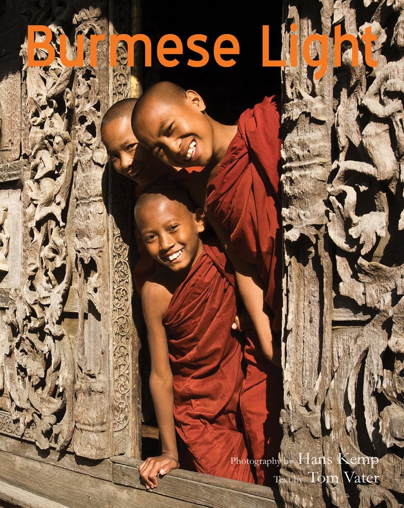 3 smiling child novice monks in robes, posing out of window of carved wood building, Burmese Light in orange font above.