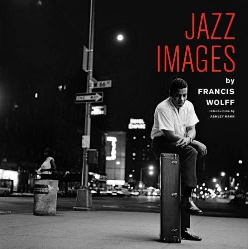 Jazz musician Wayne Shorter perched on his tenor saxophone case, on sidewalk, under night sky, JAZZ IMAGES BY FRANCIS WOLFF in red and white font to upper right.