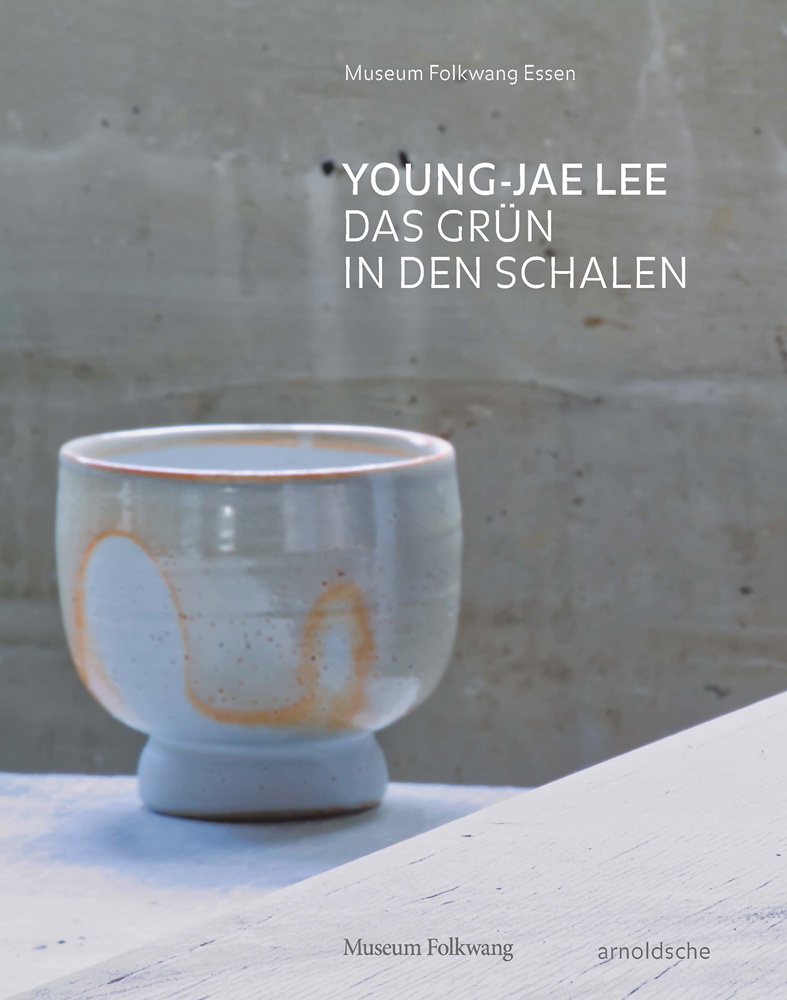 Pale blue ceramic bowl with orange markings, on white surface, YOUNG-JAE LEE DAS GRUN IN DEN SCHALEN in white font to upper right.