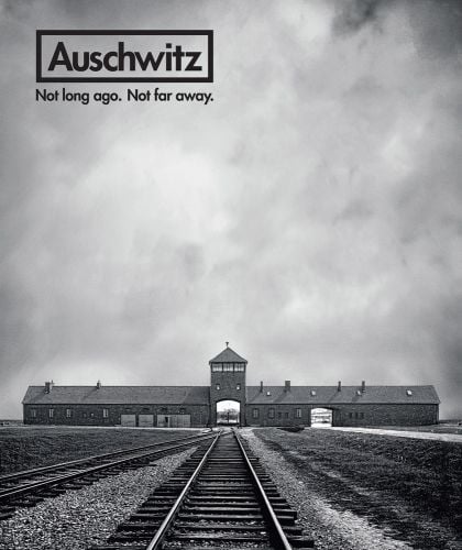 Bleak image of Auschwitz concentration camp, rail line in front, beneath cloudy sky, Auschwitz Not Long Ago. Not Far Away. in black font to top left.