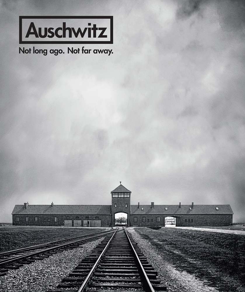 Auschwitz concentration camp, rail line in front, beneath cloudy sky, Auschwitz Not Long Ago. Not Far Away. in black font to top left.