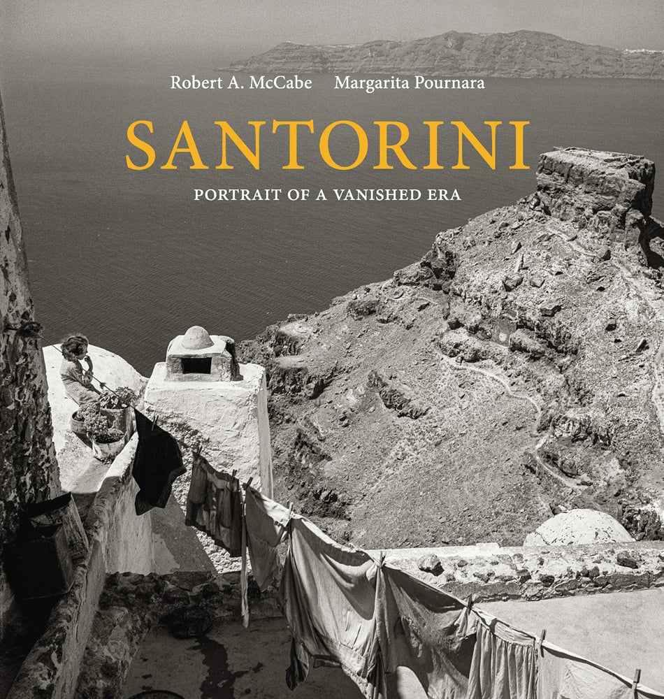 High angle shot from building looking down on washing line of clothes above rocky landscape with sea behind, SANTORINI in orange font above.