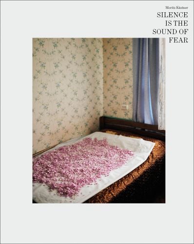 Single bed with white sheet on top, covered in pink flower petals, on white cover, Moritz Kustner SILENCE IS THE SOUND OF FEAR in black font to top right.