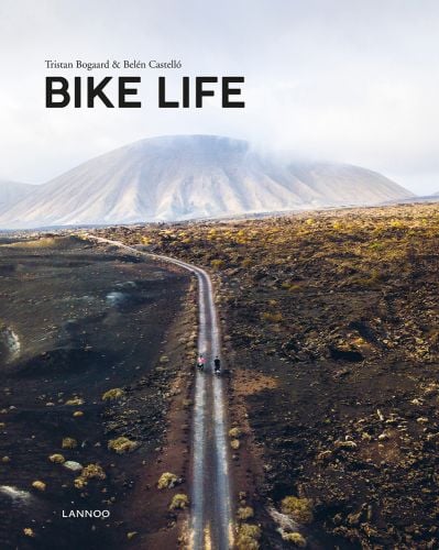 Aerial view of 2 people on bicycles riding on road towards mountainous landscape, on cover of 'Bike Life, Travel, Different', by Lannoo Publishers.
