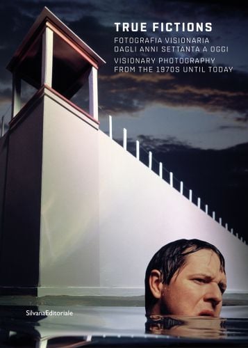 Film still of man, chin deep in water, white building with tower looming over him, True Fictions in white font to upper right.