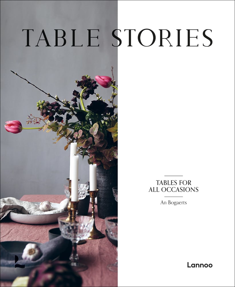 Wood table with vase of flowers, candlesticks, glasses and bowls, on cover of 'Table Stories, Tables for All Occasions', by Lannoo Publishers.