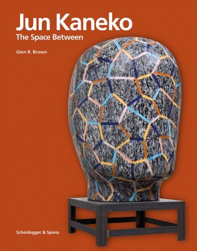 Ceramic glazed sculpture of head on low table, orange cover, Jun Kaneko The Space Between in white font to upper left.