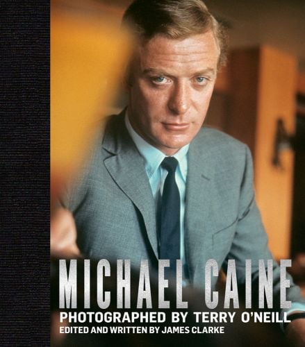Michael Caine stares at the camera wearing a grey suit with blue shirt and tie, taken from Funeral in Berlin, with silver font Michael Caine Photographed by Terry O'Neill