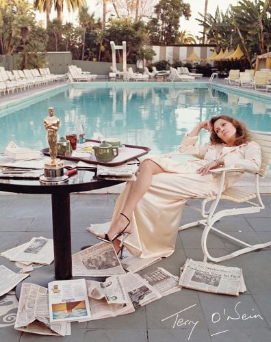 American actress Faye Dunaway lounging in a chair next to swimming pool, Oscar award on table, Terry O’Neill's printed signature in white to bottom right corner.