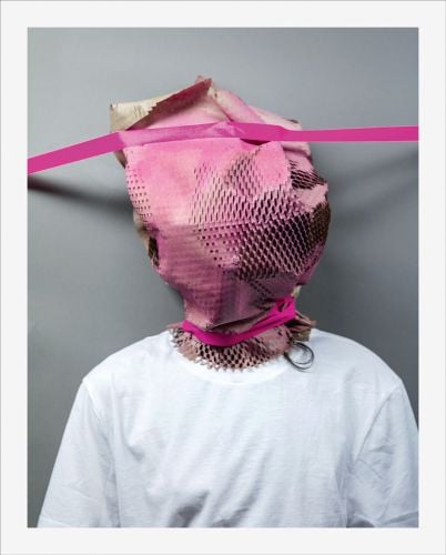 Person in white t shirt with brown shredded paper bag over head sprayed pink with pink tape across head and neck