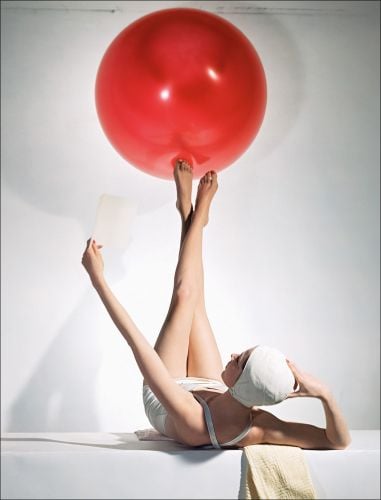 Model in white swimwear, legs in air, balancing red ball on feet, on white cover