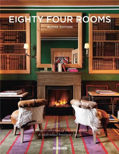 Ski resort interior with roaring fire, two fur chairs in front, 'EIGHTY FOUR ROOMS', in white font above, by teNeues Books.