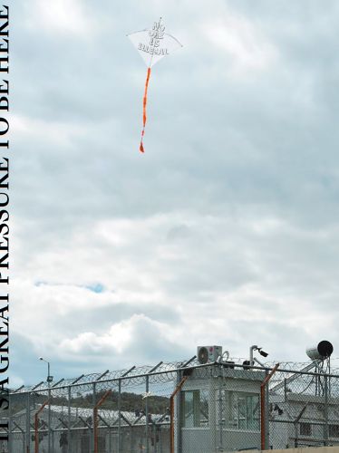 Kite flying high in sky over razor wired fence, buildings, It's a great pressure to be here in black font, top half cut off to left edge.