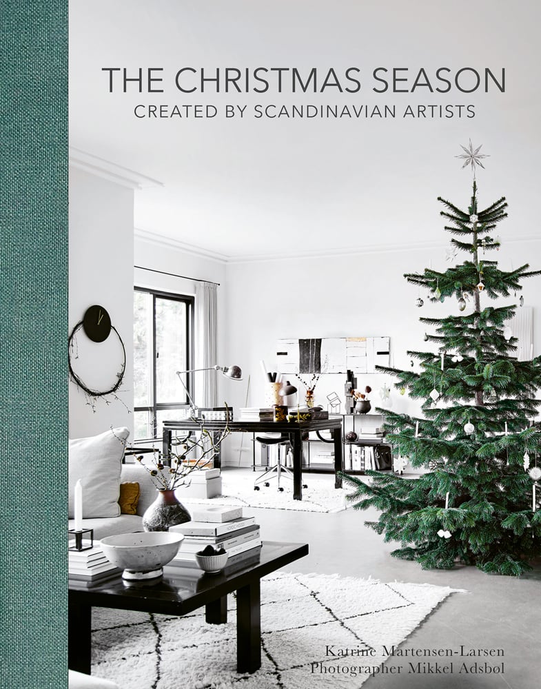 Colour photograph of a green and white festive interior with two tables and a large green fir tree with decorations and The Christmas Season Created By Scandinavian Artists in grey