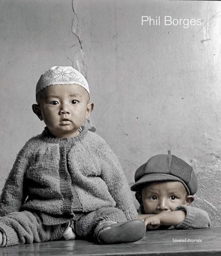 Two small children, one seated on table and one peering over, in grey woollen clothes and white cap with Phil Borges in white font above