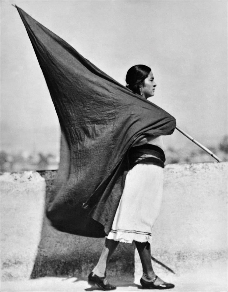 Black and white full length profile photo of woman in white dress carrying large dark fabric flag on pole