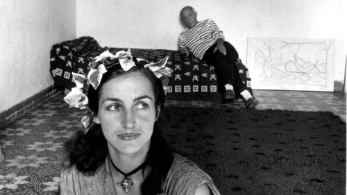 Close up of Françoise Gilot staring to her left with Pablo Picasso leaning back on bed in background