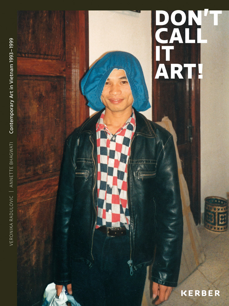 Smiling Vietnamese artist in black jacket with blue shorts on head stands in interior corridor with Don't Call it Art! in white font above