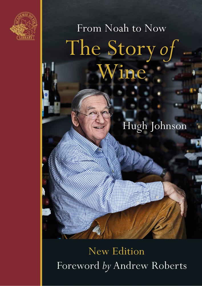 A smiling Hugh Johnson in blue checked shirt sitting inside wine store, on cover of 'The Story of Wine, From Noah to Now The Story of Wine', by Academie du Vin Library.