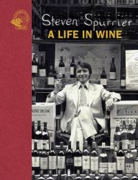 Steven Spurrier in white suit sitting in chair holding glass of wine and surrounded by stacks of wine, on cover of 'Steven Spurrier, A Life in Wine', by Academie du Vin Library.