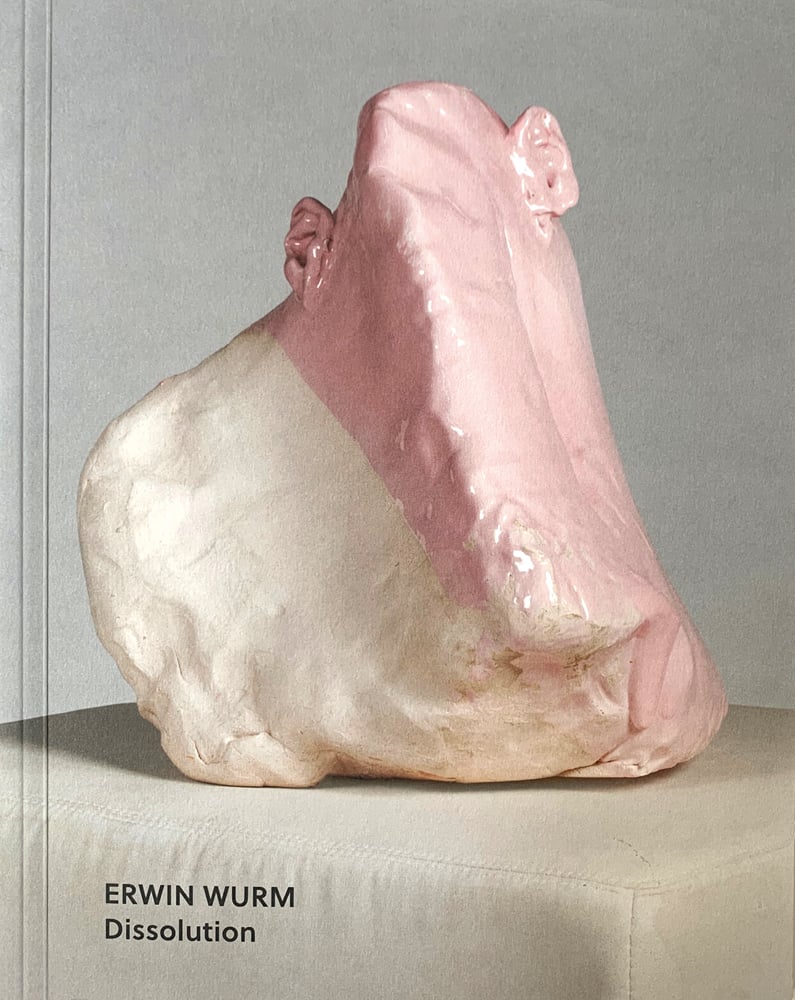 Deformed head like sculpture with ears glazed in white and pink sitting on fabric covered box, on cover of 'Erwin Wurm, Dissolution', by Arnoldsche Art Publishers.