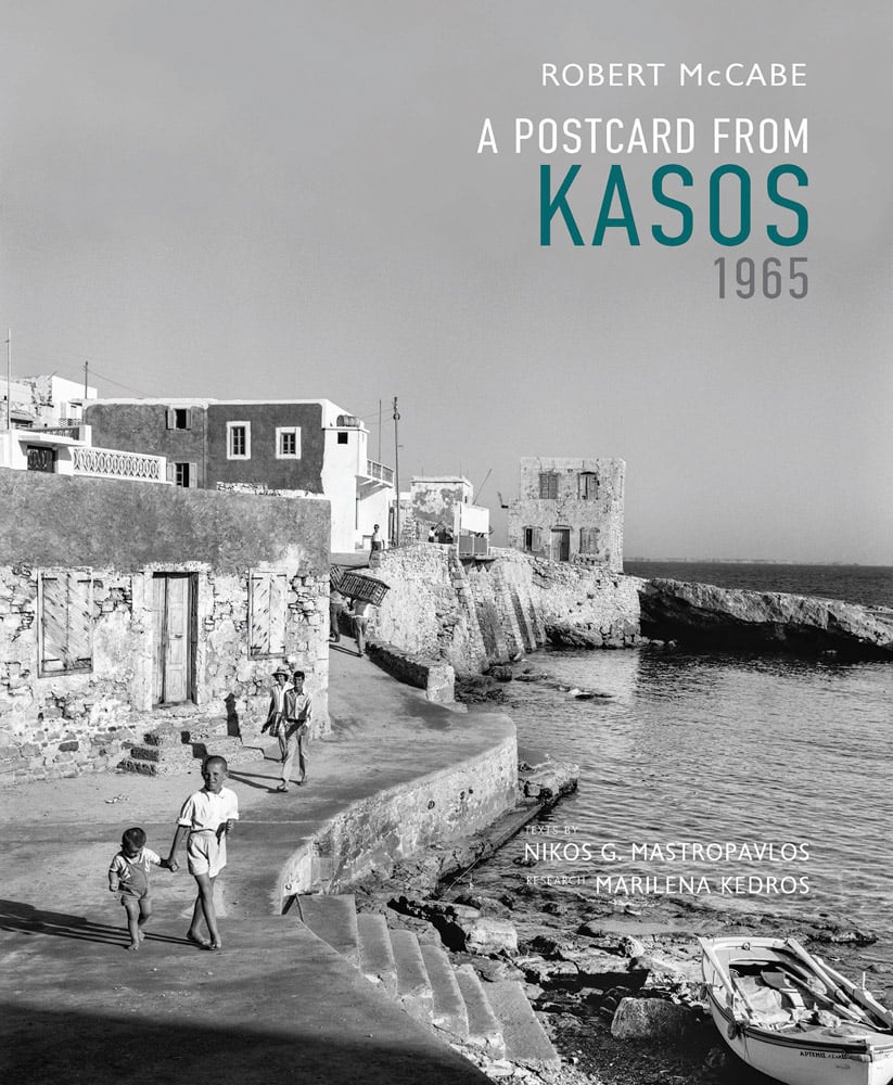 Book cover of Robert A. McCabe A Postcard from Kasos, 1965, featuring Kasos coastal area, with people walking down path next to stone buildings. Published by Abbeville Press.