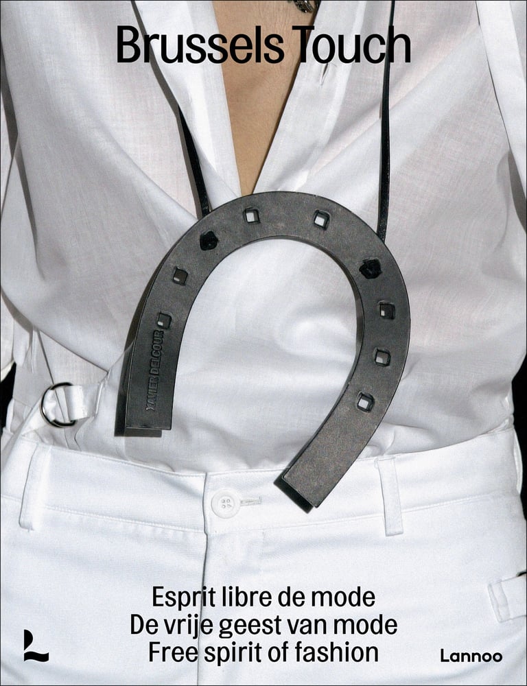 Close up of torso of model in white shirt and trousers with large horseshoe hanging around neck and Brussels Touch in black font above