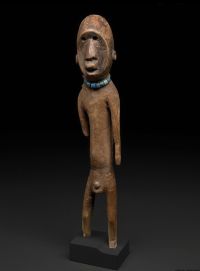 Full length brown wooden carved statue of unclothed figure on black plinth wearing a blue beaded necklace on a black background