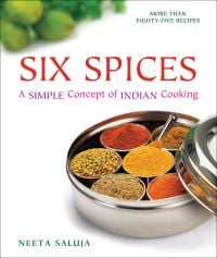 Round tin with six small dishes inside filled with red and orange herbs and spices on white surface with blurred out green fruits in background and Six Spices A Simple Concept of Indian Cooking above