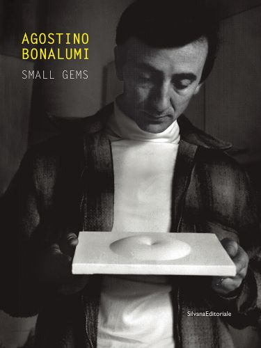 Agostino Bonalumi holding a sculptural tile with raised circular shape to centre, AGOSTINO BONALUMI SMALL GEMS in yellow and white font to upper left.